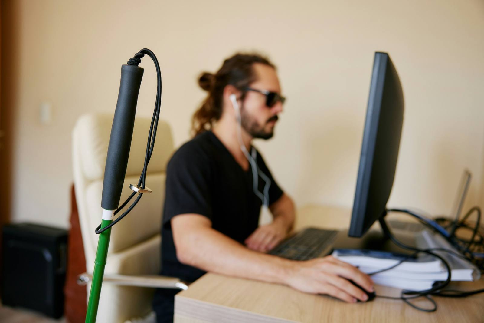 A man wearing earbuds and sunglasses works at a desktop computer, with a white cane in the foreground.